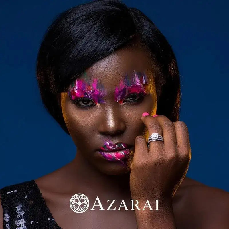 A person with artistic pink and white makeup poses against a dark blue background, wearing a ring from the Eva sterling silver bridal set and a sequined top. "Azarai" is displayed below.